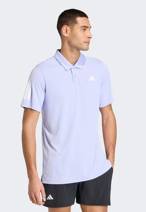 Uomo che indossa una polo Adidas lilla chiaro e pantaloncini neri Adidas, in piedi con una mano in tasca, guarda di lato.