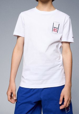 Person iført hvid Tommy Hilfiger T-shirt med "1985" logo på brystet og blå shorts mod en ens grå baggrund.