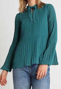 Blusa plissettata color teal con fiocco al collo, maniche lunghe e una vestibilità fluida, abbinata a jeans blu. Il tessuto ha una texture liscia e leggera.