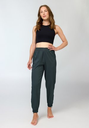 Žena stoji bosa, nosi crni bezrukavi kratki top i tamnozelene hlače za jogu na svijetlosivoj pozadini.