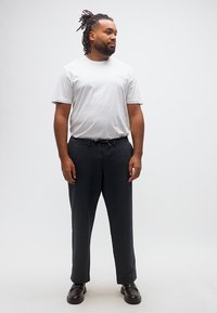 Άνδρας με dreadlocks, ντυμένος με λευκό μπλουζάκι, μαύρο παντελόνι και σκούρα καφέ παπούτσια, στέκεται μπροστά σε ένα απλό φωτεινό φόντο.