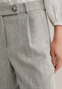 Prim-plan cu pantaloni bej cu model în țesătură herringbone, talie cu nasturi, lângă o mână și o mânecă de bluză albă texturată.