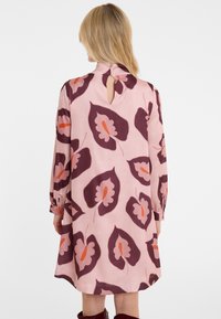 Blonde Frau in einem pinkfarbenen Kleid mit großen lila und orangefarbenen Blumenmustern, mit dem Rücken zur Kamera stehend.