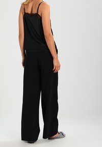 Schwarzes Tanktop mit Spitze am Saum, kombiniert mit lockeren schwarzen Hosen aus einem weichen Stoff. Die Hausschuhe haben ein graues geometrisches Muster.