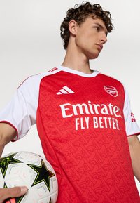 Rode en witte voetbaltop met textuurpatroon, voorzien van het logo "Emirates Fly Better" en Adidas-strepen, gecombineerd met een zwart-witte voetbal.