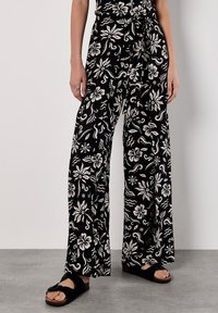 Pantaloni neri a gamba larga con motivo floreale bianco, caratterizzati da una vita alta e una cintura in tessuto, abbinati a sandali neri a scivolo su un pavimento grigio.