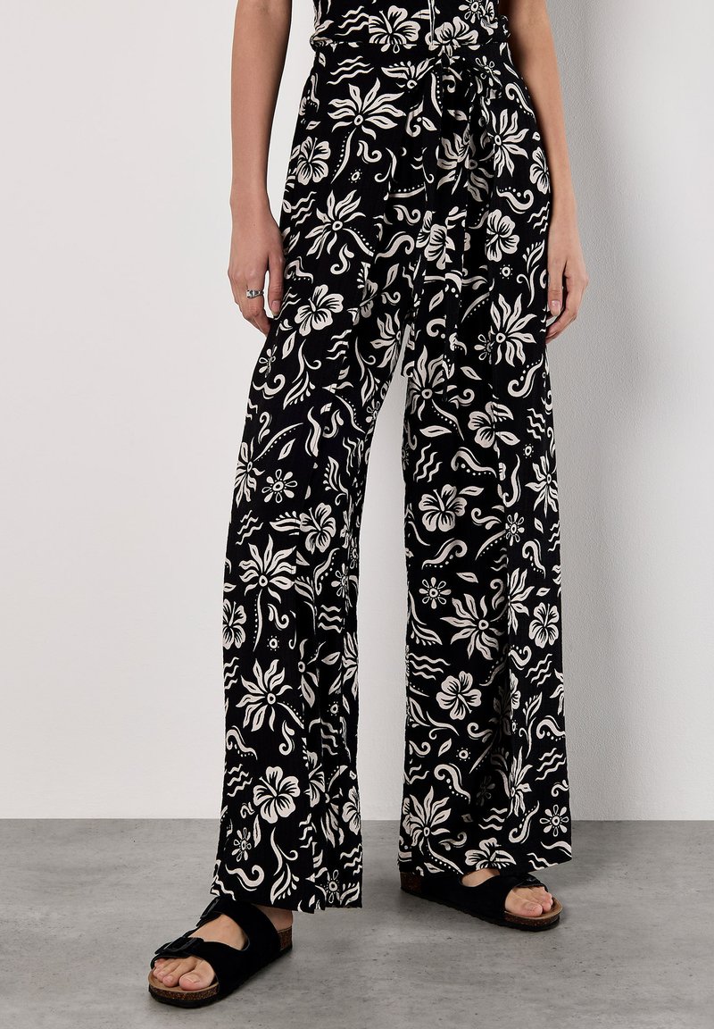 Pantaloni neri a gamba larga con motivo floreale bianco, caratterizzati da una vita alta e una cintura in tessuto, abbinati a sandali neri a scivolo su un pavimento grigio.