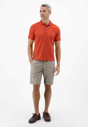 Mand stående iført en orange kortærmet polo, beige knælange shorts med snøre og brune snørestøvler, kigger mod venstre.
