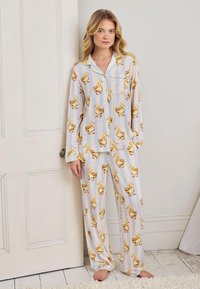 Nuori nainen seisoo paljain jaloin vaaleansinisissä raidallisissa yöpuvuissa, joissa on keltaisia ankkaprinttejä, nojaa valkoiseen oveen kirkkaassa huoneessa.