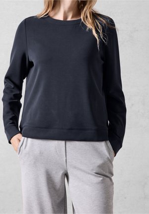 Mujer con camiseta negra de manga larga y pantalones gris claro, con las manos en los bolsillos, sobre un fondo liso gris claro.