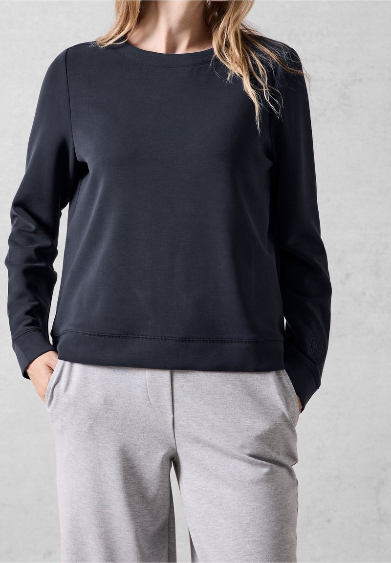 Femme portant une chemise noire à manches longues et un pantalon gris clair, les mains dans les poches, sur un fond gris clair uni.