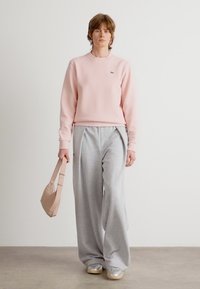 Ljusrosa sweatshirt, grå vida sweatpants och glänsande silvriga sneakers. Modellen håller en beige axelväska. Enkelt mönster, mjuk textur.