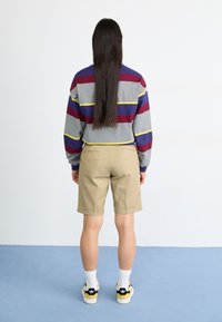 Un pull rayé gris et marron avec des accents jaunes, un short en kaki et des baskets noires avec des détails jaunes. Cheveux longs et raides.