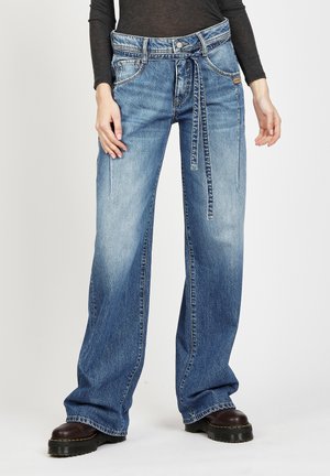 Weite, ausgewaschene blaue Jeans mit gebundenem Taillengürtel, getragen mit schwarzen klobigen Stiefeln und einem dunklen Langarmshirt.