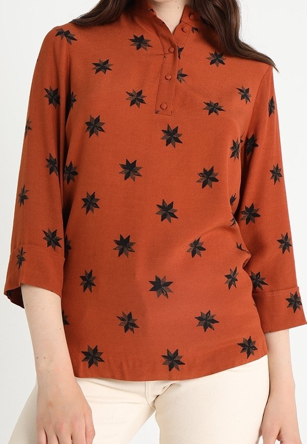 Blouse à manches longues couleur rouille avec des motifs d'étoiles noires, col boutonné, portée avec un pantalon beige clair.