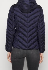 Kvinna i en marinblå vadderad jacka med huva och diagonala sömmar, tillsammans med mörkgrå åtsittande jeans, visad från baksidan.