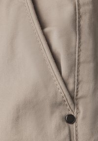 Beige tyg med synliga diagonala sömmar och en svart tryckknapp som detalj. Texturen verkar slät med en lätt glans.