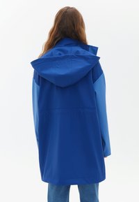 Giacca impermeabile blu con cappuccio, caratterizzata da un design bicolore, texture liscia e vita arricciata. La vista posteriore mostra una vestibilità rilassata.