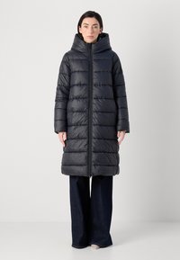 Een vrouw in een zwarte gepufferde jas met een capuchon staat rechtop, draagt een broek met wijde pijpen en schoenen in neutrale kleuren voor een effen achtergrond.