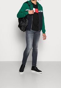 Felpa con zip verde e blu navy con strisce bianche, T-shirt nera, jeans skinny grigi e sneakers nere con suole bianche; portando uno zaino nero.