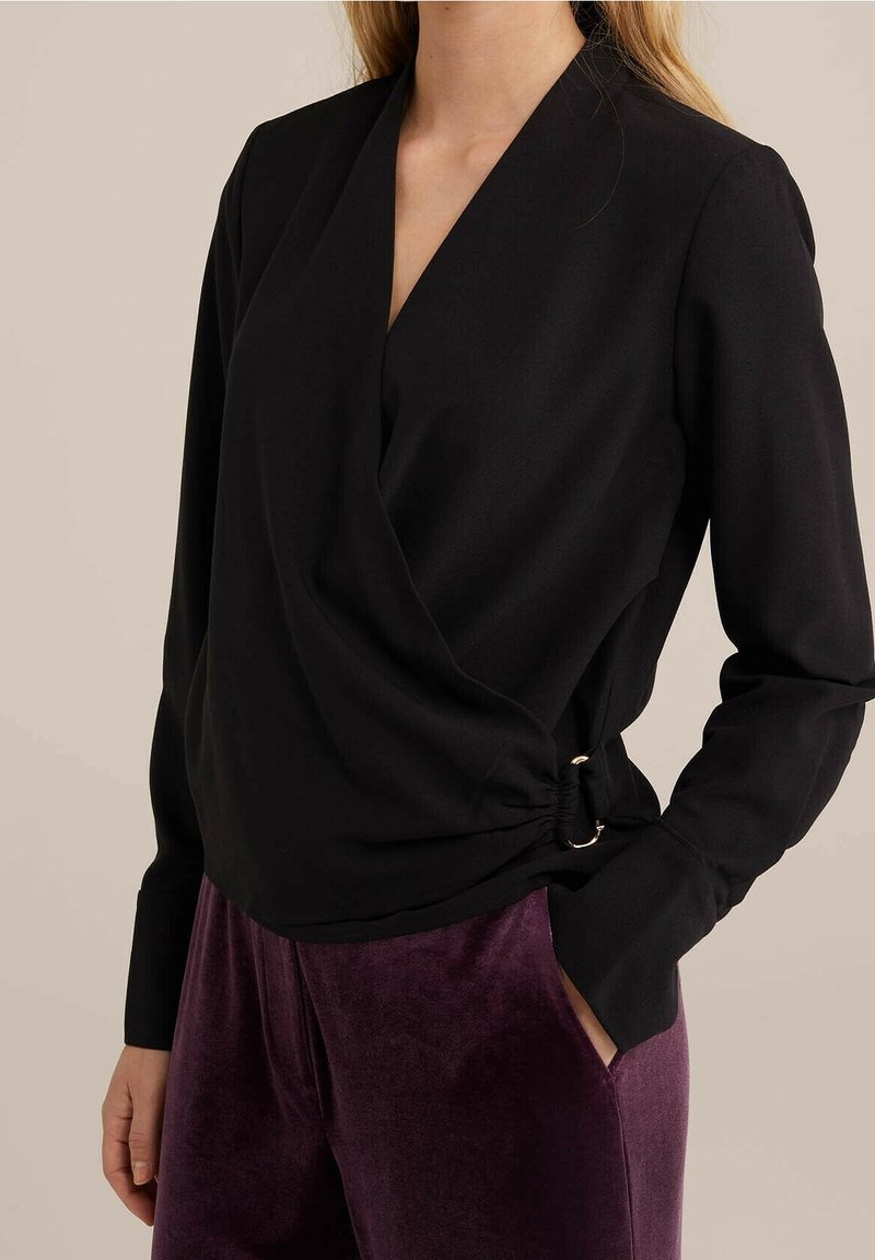 Femme portant un haut cache-cœur noir à manches longues avec un détail de boucle latérale et un pantalon en velours violet, main dans la poche sur un fond uni.