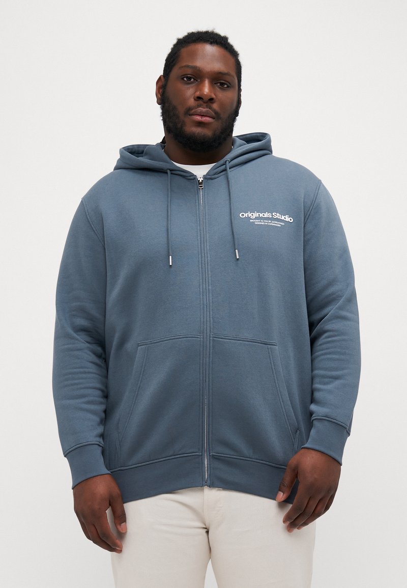 Zip-up hoodie in blue fabric, featuring a kangaroo pocket, drawstrings, and printed "Originals Studio" text on the left chest. Soft texture.