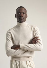 Hombre de piel oscura con un suéter de cuello alto crema y pantalones claros, de pie con los brazos cruzados contra un fondo liso.