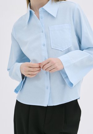 Personne portant une chemise bleu clair à manches larges, boutonnant les boutons, associée à un pantalon noir.