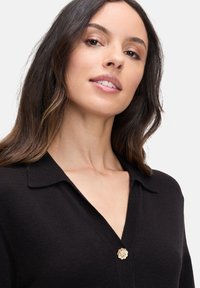 Zwarte cardigan met een V-hals, gemaakt van zachte stof. Heeft een gouden bloemenknop bij de kraag en een gladde, textuurachtige afwerking.