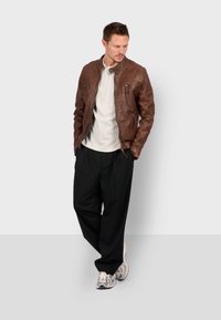 Giacca di pelle marrone con tasche con zip, colletto alto, finitura texture; abbinata a una camicia chiara e pantaloni neri larghi.