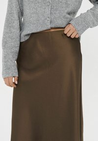Frau in einem grauen Knopf-Pullover und einem hochgeschnittenen braunen Satinrock, die ihre Rocktaille anpasst.