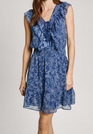 Niebieska, bezrękawnikowa sukienka w kwiaty z falbanami na dekolcie w kształcie litery V i talią podkreśloną paskiem, noszona przez osobę na jednolitym tle.