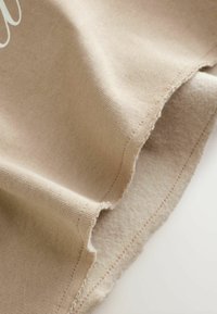 Beige bomullstyg vikt, som visar en slät textur med en ofärdig kant. Sömmen synlig längs fållen. Mjuk och lätt utseende.