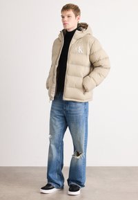Beige puffer jacket with a hood, featuring a front logo. Paired with light blue distressed jeans and black sneakers with white soles.