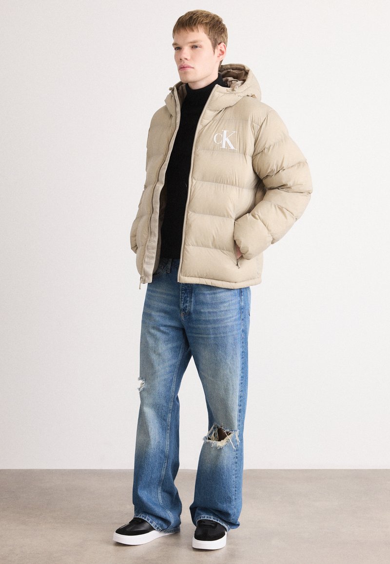 Doudoune beige avec capuche, arborant un logo sur le devant. Associée à un jean délavé bleu clair et des baskets noires avec des semelles blanches.