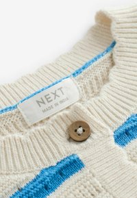 Flødefarvet strikket sweaterkrave med blå striber, en brun knap og en hvid stoflabel med teksten NEXT fremstillet i Indien.