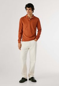 Poloko s dlhým rukávom v ohnivej oranžovej farbe, má klasický golier a dvojité zapínanie, skombinované s off-white nohavicami a čiernymi topánkami.