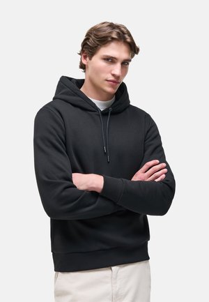 Giovane con capelli castano chiaro che indossa una felpa nera con cappuccio e pantaloni chiari, in piedi con le braccia incrociate su uno sfondo semplice.