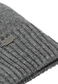 Grijze gebreide beanie met een geribbelde textuur, voorzien van een klein metalen logoaccent. De stof lijkt zacht en warm.