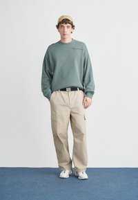 Sudadera verde con textura acanalada, pantalones cargo beige con bolsillos, zapatillas blancas y gorra beige. Diseño sencillo, ajuste casual, detalles minimalistas.