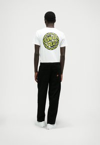 Baltā t-kreklā aizmugurē redzams melns un dzeltens apļveida grafiskais attēls. Kombinēts ar melnām biksēm un baltām sneakeru kurpēm.