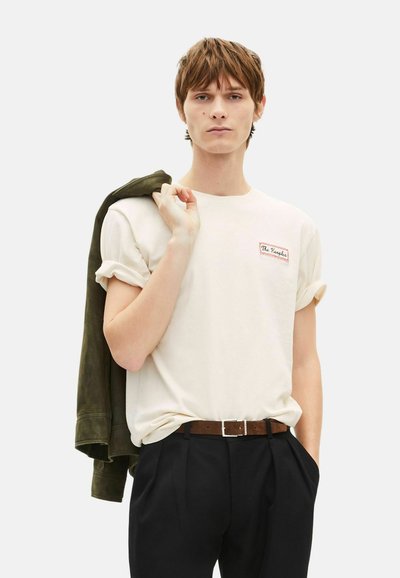 Beżowy bawełniany t-shirt z podwiniętymi rękawami, z małą czerwoną i białą naszywką z logo; w zestawie z czarnymi spodniami i oliwkowo-zieloną kurtką.