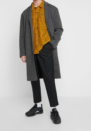 Man i grå överrock, senapsgul leopardmönstrad skjorta, svarta randiga byxor, vita strumpor och svarta sneakers, står med en hand i fickan.