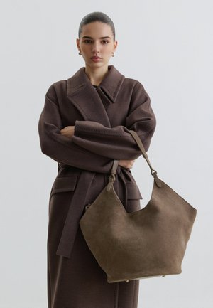 Tânără cu părul prins în spate poartă o haină maro de lână și ține o geantă mare maro din piele întoarsă pe un fundal neutru.