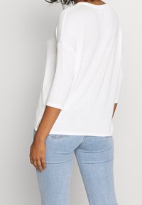 Kvinna i en lös vit tröja med trekvartsärmar och ljusblå denimjeans, sedd bakifrån.