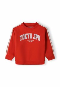 Sarkans krekls ar apaļu kakla izgriezumu, balts teksts "TOKYO JPN" un kontrastējošas sānu svītras. Mīksts audums, ribotās piedurknes un apakšmala.