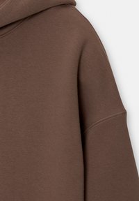 Sweat à capuche marron présentant une texture douce, une poche kangourou et des poignets côtelés. Le tissu semble lisse avec un léger éclat.