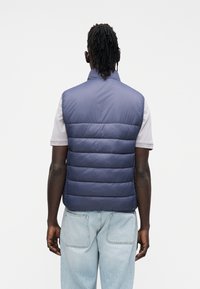 Vestă puffer bleumarin, fără mâneci, cu secțiuni matlasate orizontale. Textură moale, guler înalt, văzută din spate, asortată cu blugi de culoare albastru deschis.