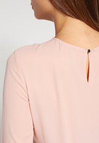 Femme aux cheveux bruns vue de dos portant un chemisier rose clair avec une ouverture en trou de serrure et une fermeture par bouton au niveau de l'encolure.