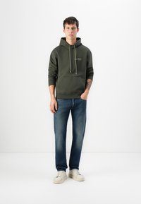 Young man standing with hands in pockets, wearing an olive green hoodie, blue jeans, white sneakers, and a visible forearm tattoo.